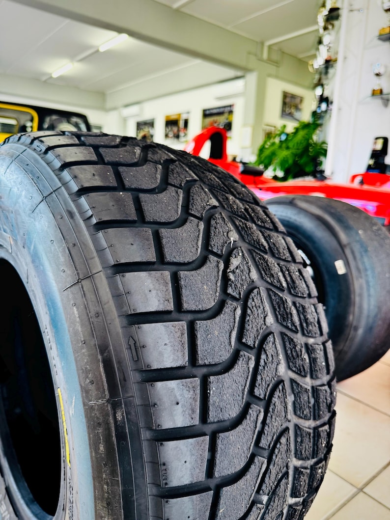 May include: A black rubber tire featuring a deep, intricate tread pattern with rectangular blocks and grooves. The tire surface shows signs of wear and texture. A small directional arrow is embossed on the sidewall, indicating rotation.