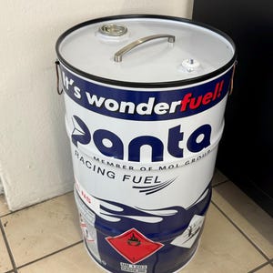 May include: A white and blue metal drum with the text "panta racing fuel" and "It's wonderfuel!" The drum has a silver handle and a red and white hazard label. The drum is cylindrical.