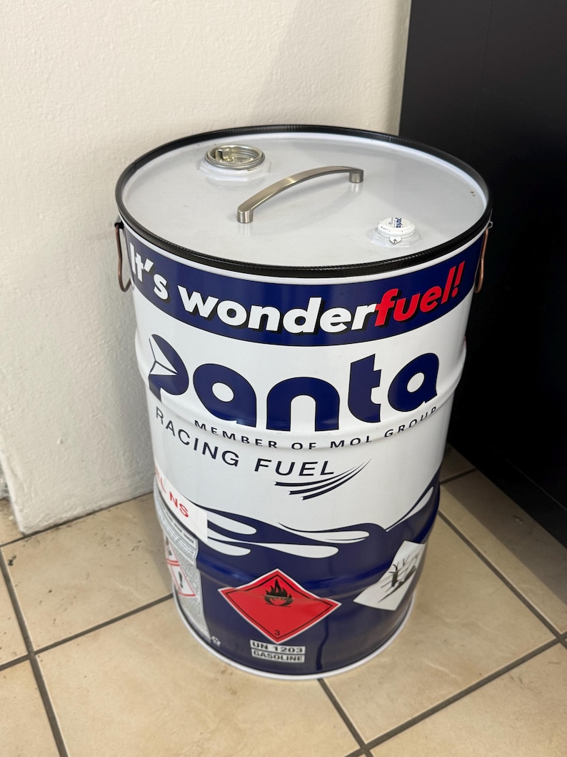 May include: A white and blue metal drum with the text "panta racing fuel" and "It's wonderfuel!" The drum has a silver handle and a red and white hazard label. The drum is cylindrical.