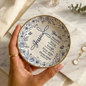Puede incluir: Un plato decorativo blanco con borde dorado, con un diseño floral azul. El plato tiene el nombre "Jessica" y las palabras "You are..." con texto inspirador. El plato es sostenido por una persona.