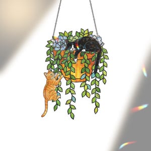 May include: A stained glass hanging decoration featuring a potted plant with a black and white cat resting on top. An orange tabby cat is climbing the pot. The plant has green leaves and blue flowers. The pot is orange and hangs from a chain.