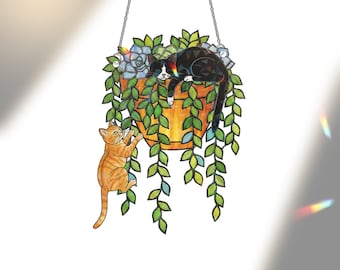 Puede incluir: Una decoración colgante de vidrieras que representa una maceta con un gato blanco y negro descansando encima. Un gato atigrado naranja trepa por la maceta. La planta tiene hojas verdes y flores azules. La maceta es naranja y cuelga de una cadena.