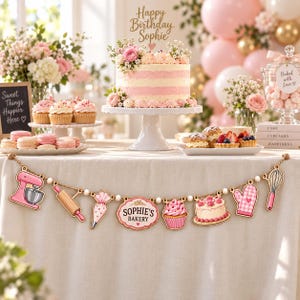 May include: A decorative garland featuring wooden cutouts of a pink stand mixer, rolling pin, piping bag, a sign reading SOPHIE'S BAKERY, a cupcake, a cake, a pink oven mitt, and a whisk, all strung on a natural twine cord.