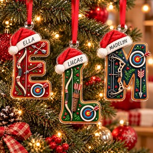 May include: Three clear acrylic letter ornaments shaped as E, L, and M, each topped with a red Santa hat. The letters feature archery motifs including bows, arrows, targets, and quivers, with the names ELLA, LUCAS, and MADELYN printed on the hats.