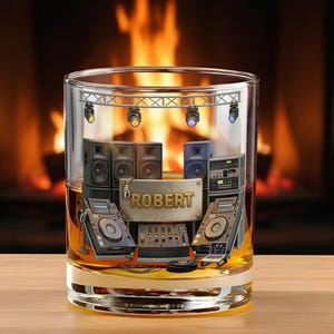 May include: A rocks glass with a stage design featuring speakers, DJ equipment, and the name "ROBERT" on a gold plaque. The glass is filled with a golden liquid, likely whiskey, and set against a blurred fireplace background.
