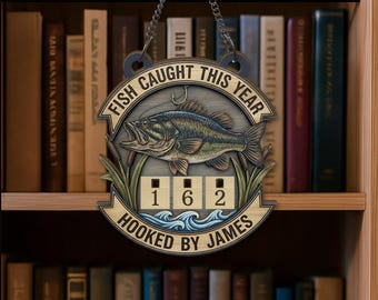 Op de afbeelding: Houten bord met een visontwerp en de tekst "FISH CAUGHT THIS YEAR". Het bord heeft een haak en een visillustratie, met de cijfers "162" en de tekst "HOOKED BY JAMES". Het hangt aan een ketting.