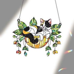 May include: Stained glass cat suncatcher. A calico cat rests in a yellow basket surrounded by green leaves and orange flowers. The cat has black, white, and orange markings. The suncatcher hangs from a silver chain.