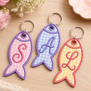 Op de afbeelding: Drie visvormige sleutelhangers met metalen ringen. De linker sleutelhanger is roze geblokt met een paarse rand en de letter S. De middelste sleutelhanger is blauw geblokt met een paarse rand en de letter A. De rechter sleutelhanger is geel geblokt met een rode rand en de letter L.