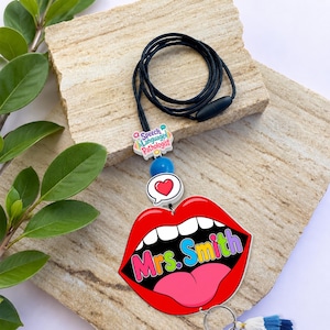 May include: A black lanyard featuring a charm with the text Speech Language Pathologist, a blue bead, a white bead with a red heart, and a large red lip-shaped pendant with the text Mrs. Smith in colourful letters and a pink tongue.