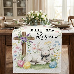 May include: White Easter table runner with a lamb, cross, and floral design. The runner features the text "HE IS Risen" and includes colorful Easter eggs. The runner is placed on a wooden table with place settings and a floral centerpiece.