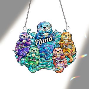Könnte beinhalten: Ein Anhängeschild im Glasmalerei-Stil mit einer Otterfamilie. Der zentrale Otter, mit der Aufschrift "Nana", ist von Ottern namens Jackson, Emily, Sophia und Molly umgeben. Jeder Otter hält eine Muschel. Das Schild hängt an zwei Ketten.