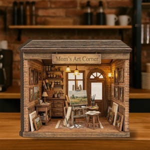 Puede incluir: Diorama en miniatura de un estudio de arte con el letrero "Mom's Art Corner". El estudio presenta una fachada de ladrillo, pinturas, un caballete con un paisaje y materiales de arte. El interior está iluminado con luces cálidas.