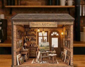 Op de afbeelding: Miniatuur diorama van een kunststudio met het bord "Mom's Art Corner". De studio heeft een bakstenen gevel, schilderijen, een ezel met een landschap en kunstbenodigdheden. Het interieur wordt verlicht door warm licht.