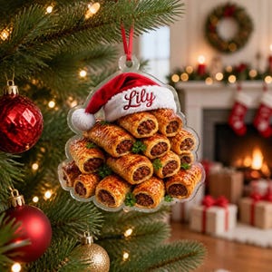 May include: A clear acrylic Christmas ornament shaped like a stack of sausage rolls, topped with a red and white Santa hat featuring the name Lily embroidered in red script. The ornament hangs from a red ribbon and displays golden-brown pastry.