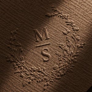 Peut inclure: Une surface en papier texturé brun foncé présentant un monogramme central en relief avec les lettres M et S séparées par une ligne horizontale, le tout entouré d'une couronne en relief détaillée de petites baies et de branches florales.