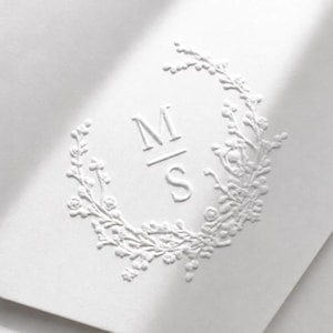 Op de afbeelding: Een witte kaart met een blinddrukontwerp van een bloemenkrans die de letters M en S omsluit, gescheiden door een horizontale lijn. De reliëfelementen steken uit boven het oppervlak en creëren een subtiele textuur en diepte op het papier.