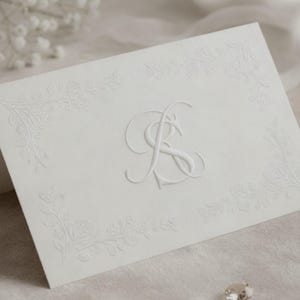 May include: A white folded card featuring embossed floral patterns and a central monogram. The card, likely a wedding invitation or stationery, is set against a soft, neutral backdrop, with delicate details.