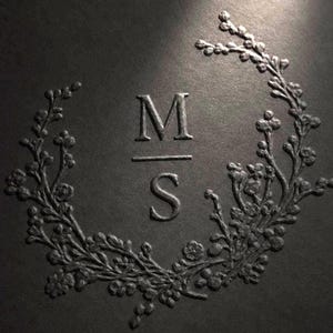 Puede incluir: Una superficie texturizada de color gris oscuro con un diseño en relieve de las letras M y S separadas por una línea horizontal, todo ello encerrado en una corona circular de pequeñas ramas florales detalladas y capullos a tono.