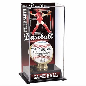 May include: A baseball game ball displayed in a clear acrylic case. The ball is white with red stitching and sits on a gold-colored base. The case includes a graphic of a baseball player and the text "GAME BALL".