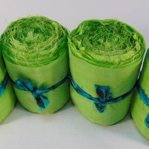 Puede incluir: Cuatro rollos de papel verde brillante, cada uno atado con una cinta turquesa. El papel tiene una apariencia texturizada y en capas, y los rollos están dispuestos uno al lado del otro sobre un fondo blanco.