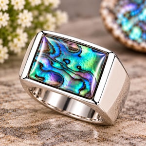 May include: A silver-toned ring featuring a rectangular inlay of iridescent abalone shell. The shell displays a vibrant mix of blue, green, and purple hues with natural swirling patterns set within a polished, smooth metal band with beveled edges.