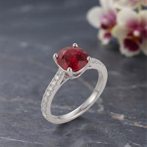 May include: A silver ring featuring a round, vibrant red gemstone set in a four-prong mounting. The band is crafted from polished silver and is accented with a row of small, clear pavé-set stones along the top half of the shank on both sides.