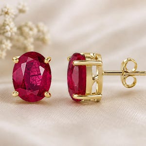 Pode incluir: Um par de brincos de tarracha com pedras preciosas vermelho-escuras de corte oval cravadas em ouro amarelo polido. Um brinco é mostrado de frente, enquanto o outro exibe o perfil lateral, a cravação de quatro garras e o pino de ouro.