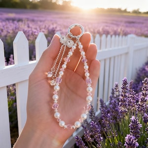 Op de afbeelding: Een kralen sleutelhanger met een zilveren sluiting, met witte parels en roze kristallen kralen. De sleutelhanger heeft een decoratieve bedel met bungelende zilveren kettingen en heldere kristallen accenten. De achtergrond toont een lavendelveld.