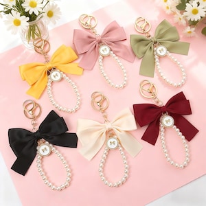 May include: A selection of keyrings with satin bows in various colours. Each keyring has a bow, a gold-tone clasp, a pearl bead strand, and a small circular charm with a bow design. Colours include yellow, pink, green, black, cream, and burgundy.