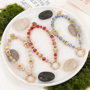 Può includere: Tre portachiavi con perline rosa, rosse e blu, ciascuno con dettagli dorati e un grande anello dorato. I portachiavi presentano perline e ciondoli decorativi, tra cui gufi, farfalle e forme geometriche. I portachiavi sono esposti su un piatto bianco con pietre decorative.