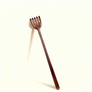 May include: A dark brown wooden back scratcher with a long handle. The scratching head has five tines. The handle is slender and ends in a rounded shape. The back scratcher is set against a white background.