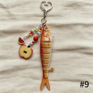 Puede incluir: Un llavero decorativo con un dise&ntilde;o de pez segmentado en naranja y amarillo, con un cierre en forma de coraz&oacute;n. Incluye un dije con una manzana y una sand&iacute;a, y un acento de cuentas. El n&uacute;mero "#9" es visible.