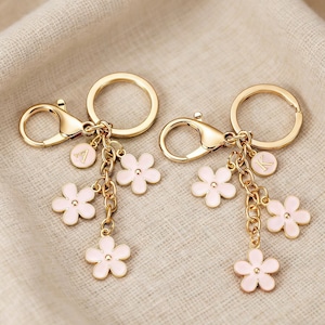 May include: Two gold-colored keychains with a lobster clasp and a round key ring. Each keychain has a chain with three pink flower charms and a small round charm with a letter. The background is a light beige fabric.