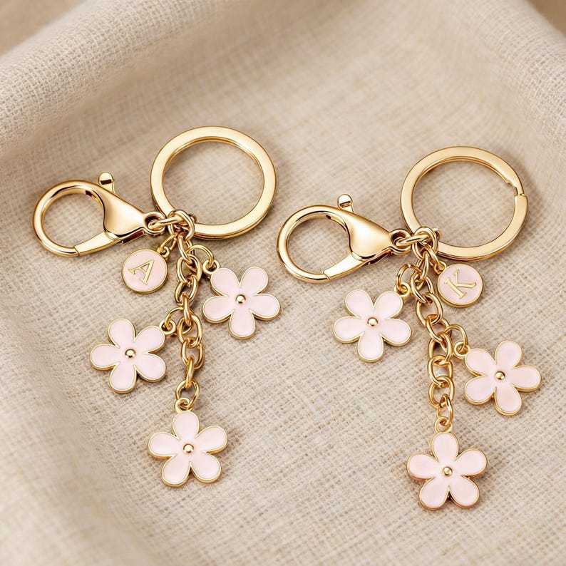 May include: Two gold-colored keychains with a lobster clasp and a round key ring. Each keychain has a chain with three pink flower charms and a small round charm with a letter. The background is a light beige fabric.