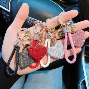 Op de afbeelding: Een verzameling hartvormige sleutelhangers met strass-versieringen. Elke sleutelhanger heeft een hart in een andere kleur, waaronder zwart, rood, wit en roze. De sleutelhangers hebben goudkleurige hardware en gevlochten leren bandjes.