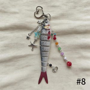 Puede incluir: Un llavero con forma de pez con un cierre en forma de coraz&oacute;n. El pez es blanco y gris con detalles rojos y una cola roja. Est&aacute; decorado con abalorios de estrellas y cuentas, y el n&uacute;mero "#8" est&aacute; en la esquina inferior derecha.