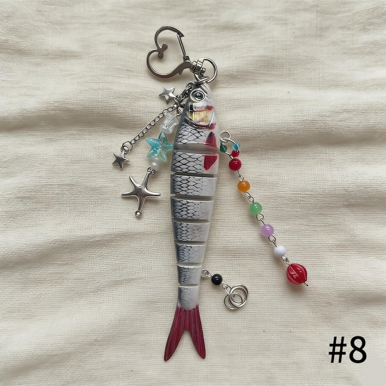 Puede incluir: Un llavero con forma de pez con un cierre en forma de coraz&oacute;n. El pez es blanco y gris con detalles rojos y una cola roja. Est&aacute; decorado con abalorios de estrellas y cuentas, y el n&uacute;mero "#8" est&aacute; en la esquina inferior derecha.