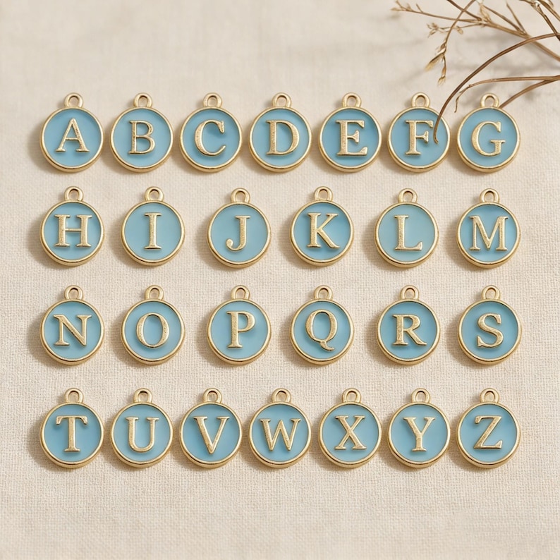 May include: A collection of gold-colored alphabet charms, each featuring a letter from A to Z. The letters are in a gold font, set against a light blue enamel background, and surrounded by a gold border. Each charm has a small loop at the top.