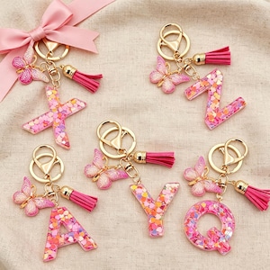May include: Five keychains featuring resin letters X, Z, A, Y, and Q filled with pink and orange heart-shaped glitter. Each keychain includes a gold-tone metal ring, a pink faux leather tassel, and a pink butterfly charm with glitter accents.