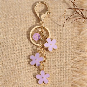 May include: Gold-colored keychain with a lobster clasp, a circular charm with the letter "A", and three lavender flower charms. The flowers have a gold outline and are connected by a delicate chain. The keychain is a decorative accessory.