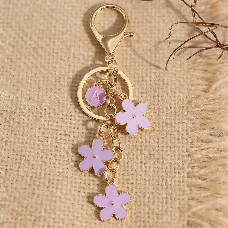 May include: Gold-colored keychain with a lobster clasp, a circular charm with the letter "A", and three lavender flower charms. The flowers have a gold outline and are connected by a delicate chain. The keychain is a decorative accessory.