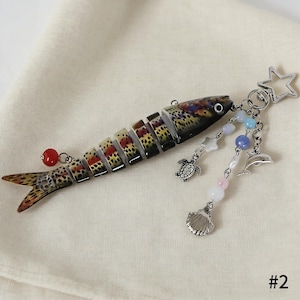 Puede incluir: Un llavero con forma de pez con un cuerpo segmentado, que presenta un colorido patr&oacute;n de manchas marrones, rojas y amarillas. Tiene un charm de cuentas rojas y un charm colgante con una estrella, una tortuga, una concha y un delf&iacute;n.