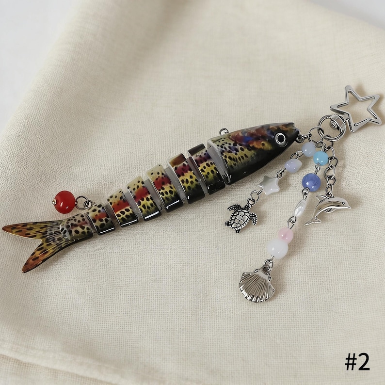 Puede incluir: Un llavero con forma de pez con un cuerpo segmentado, que presenta un colorido patr&oacute;n de manchas marrones, rojas y amarillas. Tiene un charm de cuentas rojas y un charm colgante con una estrella, una tortuga, una concha y un delf&iacute;n.
