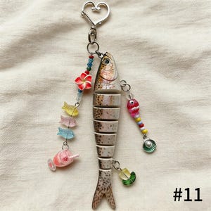 Puede incluir: Un llavero decorativo con forma de pez con un cierre plateado en forma de coraz&oacute;n. El pez est&aacute; segmentado con detalles marrones y blancos, adornado con cuentas de colores, dijes de peces en miniatura y una flor roja. El n&uacute;mero "#11" est&aacute; impreso en la esquina inferior derecha.
