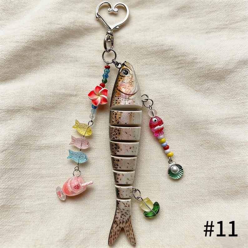 Puede incluir: Un llavero decorativo con forma de pez con un cierre plateado en forma de coraz&oacute;n. El pez est&aacute; segmentado con detalles marrones y blancos, adornado con cuentas de colores, dijes de peces en miniatura y una flor roja. El n&uacute;mero "#11" est&aacute; impreso en la esquina inferior derecha.