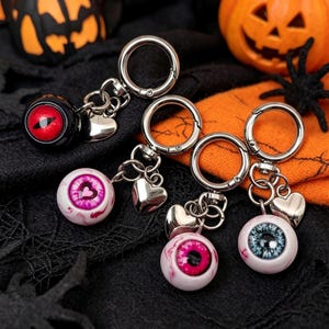 Pode incluir: Quatro porta-chaves de metal prateado, cada um com um pendente de globo ocular esférico e um pequeno pendente em forma de coração. Os globos oculares têm íris distintas em vermelho, rosa com pupila de coração, rosa vivo e azul, em argolas.