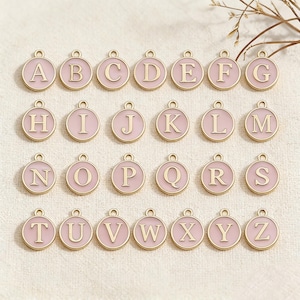 May include: A collection of twenty-six round, pink enamel alphabet charms with gold-colored borders and capital letters. Each charm has a small loop at the top for attaching to a chain or bracelet. The letters A through Z are displayed.