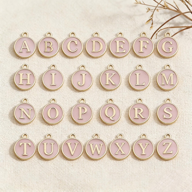 May include: A collection of twenty-six round, pink enamel alphabet charms with gold-colored borders and capital letters. Each charm has a small loop at the top for attaching to a chain or bracelet. The letters A through Z are displayed.