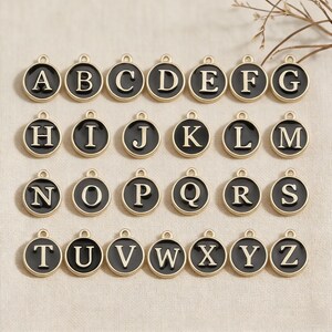 May include: A collection of gold-toned alphabet charms, each featuring a black enamel background and a capital letter in a serif font. The charms are round with a small loop at the top for attaching to a chain or bracelet.
