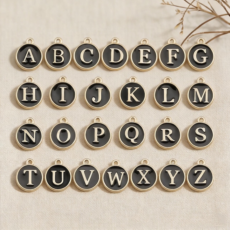 May include: A collection of gold-toned alphabet charms, each featuring a black enamel background and a capital letter in a serif font. The charms are round with a small loop at the top for attaching to a chain or bracelet.